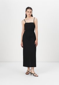 Robe noire sans manches avec décolleté carré, silhouette slim et longueur au genou. Modèle portant des talons noirs à lanières. Texture de tissu lisse.