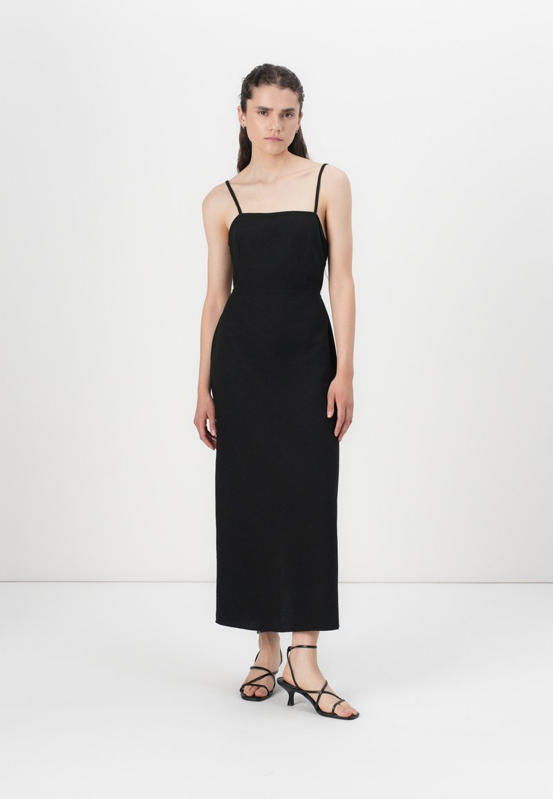 Robe noire sans manches avec décolleté carré, silhouette slim et longueur au genou. Modèle portant des talons noirs à lanières. Texture de tissu lisse.
