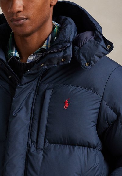 Tengerészkék pehelydzseki magas gallérral, nagy első zsebbel és piros polo logóval a mellrészen. Simább, fényes felület.