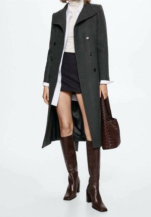 Mujer que lleva abrigo gris oscuro de doble botonadura, cuello alto blanco, minifalda negra, botas marrones hasta la rodilla, sosteniendo un bolso marrón tejido.