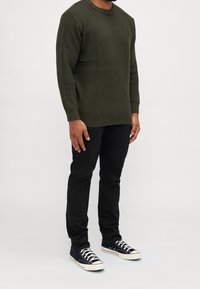 Man med en olivgrön stickad tröja, svarta snäva byxor och svarta låga sneakers med vita snöre och sulor, står mot en enkel bakgrund.