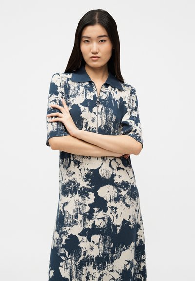 Γυναίκα με μακριά σκούρα μαλλιά, που φοράει φόρεμα με μπλε και μπεζ αφηρημένο σχέδιο, γιακά και κοντά μανίκια, στεκόμενη με σταυρωμένα τα χέρια.