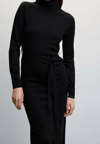 Robe à col roulé noire en tissu doux, avec manches longues, silhouette ajustée et ceinture à nouer à la taille pour un ajustement modulable.