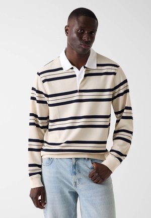 Młody mężczyzna w beżowo-granatowej koszulce rugby z długim rękawem, białym kołnierzykiem i jasnoniebieskich dżinsach, stojący na tle gładkiej ściany.