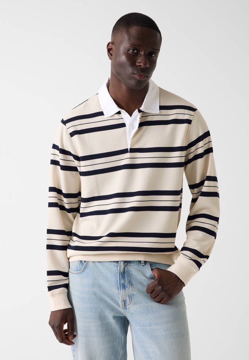 Jeune homme portant un polo rugby à manches longues beige et bleu marine à rayures avec un col blanc, et un jean bleu clair, debout devant un fond uni.
