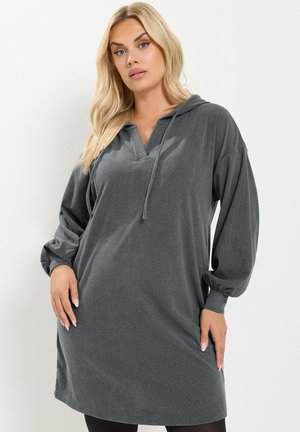 Femme blonde aux cheveux lâchés, portant une robe à capuche gris foncé à manches longues avec un col en V et des cordons, debout devant un fond uni.