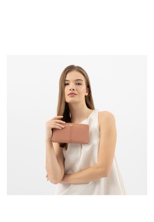 Jonge vrouw in een wit mouwloos topje houdt een beige portemonnee met rits dicht bij haar gezicht tegen een effen witte achtergrond.