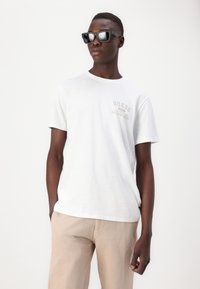 Hvit bomull T-skjorte med "Guess" logo i lys brun, sammen med lyse beige stripete bukser. Modellen har på seg svarte firkantede solbriller.