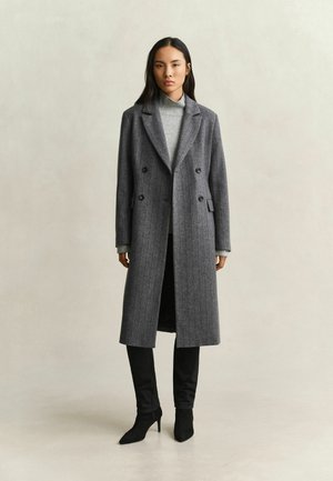Femme debout portant un long manteau gris croisé, un pull à col roulé gris clair, un pantalon noir et des bottes à talons noires sur un fond uni.