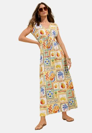 Frau trägt ein farbenfrohes Maxikleid mit Obst- und Blumenmustern, Sonnenbrille, Armbändern und braunen Sandalen, steht mit einer Hand an der Hüfte.