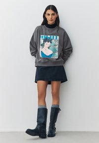 Harmaa collegepaita jossa graafinen painatus, musta hame ja polven korkuiset siniset saappaat, joissa on teksturoidut sukat. Tyylikäs rento asukokonaisuus, jossa kerroksellisia vaatekappaleita.