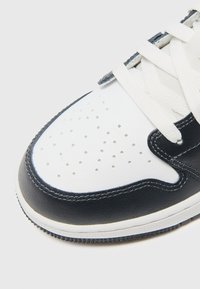 Zapatilla con parte superior de cuero blanco, acentos en azul marino, lengüeta de malla, punta redondeada, diseño perforado y cordones blancos. Suela de goma visible.