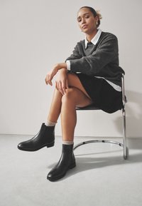 Zwarte leren enkellaarsjes met elastische zijkanten, ronde neus en lage blokhak, gedragen met grijze sokken. Model zittend met gekruiste benen in een stoel.