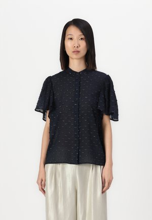 Blusa blu navy realizzata in tessuto leggero con maniche corte a volant e decorazioni testurizzate, abbinata a pantaloni chiari e lucenti.