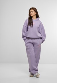 Dviejų dalių laisvalaikio kostiumas šviesiai violetinės spalvos. Sudaro laisvas džemperis su priekiu kišene ir atitinkamos tapered kelnės. Rožinės spalvos logotipo detalė.