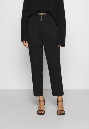 Femme portant un pantalon noir taille haute avec une ceinture à logo Pinko, associée à des sandales à talons noires et un pull noir à manches longues.