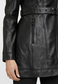 Zwarte leren jas met een tailleband, zichtbare naden en een zijvak met ritssluiting. Gladde textuur met een getailleerd ontwerp.