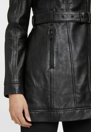 Giacca in pelle nera con vita con cintura, cuciture visibili e tasca laterale con cerniera. Texture liscia con design aderente.