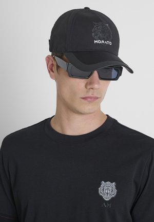 Joven con gorra negra con logo de tigre, gafas de sol rectangulares negras y camiseta negra con diseño de cara de tigre blanco en el pecho.