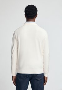 Helles Cremefarbener Pullover mit langen Ärmeln, gerippten Bündchen und Saum, weicher Textur, getragen über dunkelblauen Jeans, von hinten gesehen.