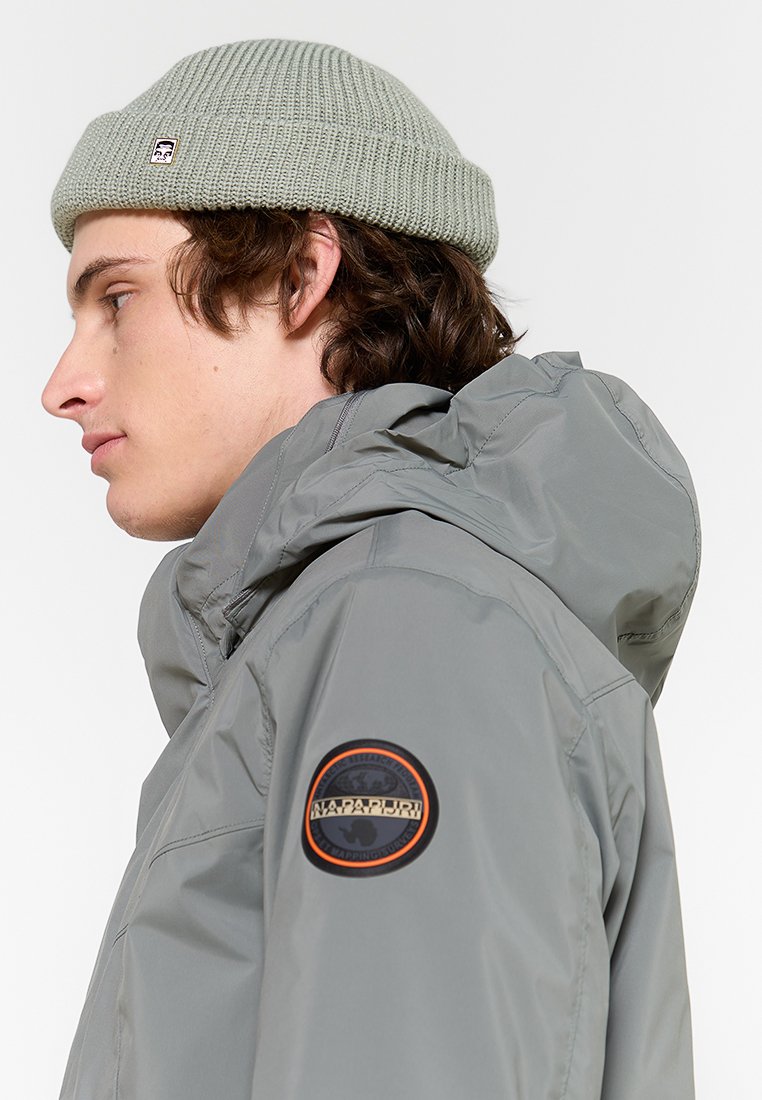 Giacca impermeabile grigia con cappuccio, dotata di un logo rotondo sul braccio sinistro e un berretto verde a maglia strutturata. Vista di profilo laterale.