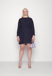 Vestido navy com mangas longas, faixa na cintura, tecido suave e um ajuste relaxado, combinado com uma pequena bolsa light purple e sandálias douradas de tiras.