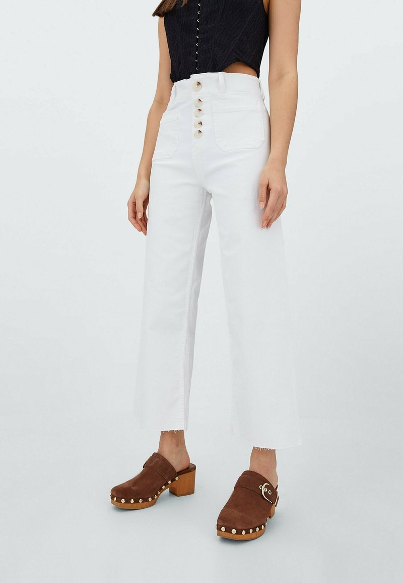 Mujer vestida con jeans de pierna ancha blancos de cintura alta con múltiples botones y zuecos marrones de tacón con tachuelas doradas sobre un fondo claro.