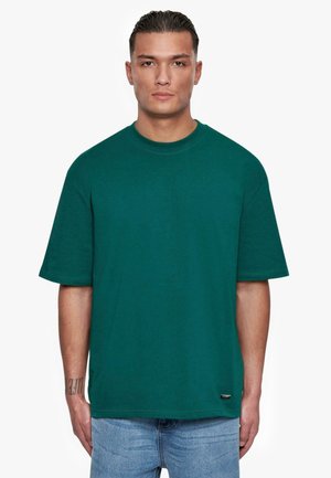 Green cotton t-shirt with short sleeves, crew neck, and relaxed fit. Features a small brand tag at the hem. Model wears blue jeans.
