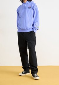 Ljuslila hoodie med framficka, grafiska mönster på bröstet och ärmen, kombinerad med svarta byxor och svarta sneakers på ett beige golv.