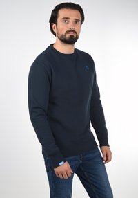 Navy blue sweatshirt in a soft material, featuring a crew neck, ribbed cuffs, and a small blue logo on the chest. Paired with blue jeans.