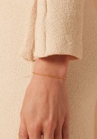 Gouden kettingarmband met een delicate gedraaide vorm, gedragen om de pols tegen een gestructureerde beige achtergrond.