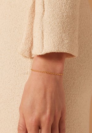 Gouden kettingarmband met een delicate gedraaide vorm, gedragen om de pols tegen een gestructureerde beige achtergrond.