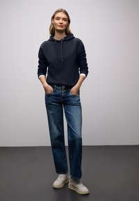 Jeune femme debout, les mains dans les poches, portant un sweat à capuche bleu marine, un jean bleu et des baskets blanches, sur un fond gris uni.