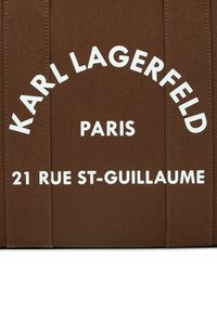 Brūna kanvas soma ar baltu tekstu: "KARL LAGERFELD PARIS 21 RUE ST-GUILLAUME," ar vertikālu šuvju, kas piešķir papildu tekstūru.