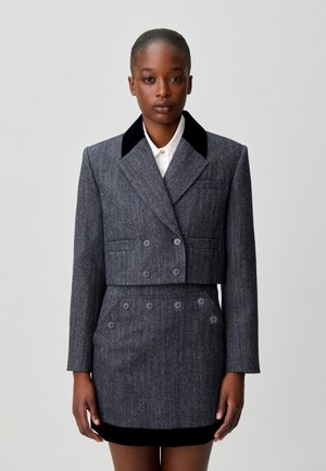 Femme portant un blazer court croisé gris foncé avec col noir et une jupe boutonnée taille haute assortie, sur une chemise blanche.