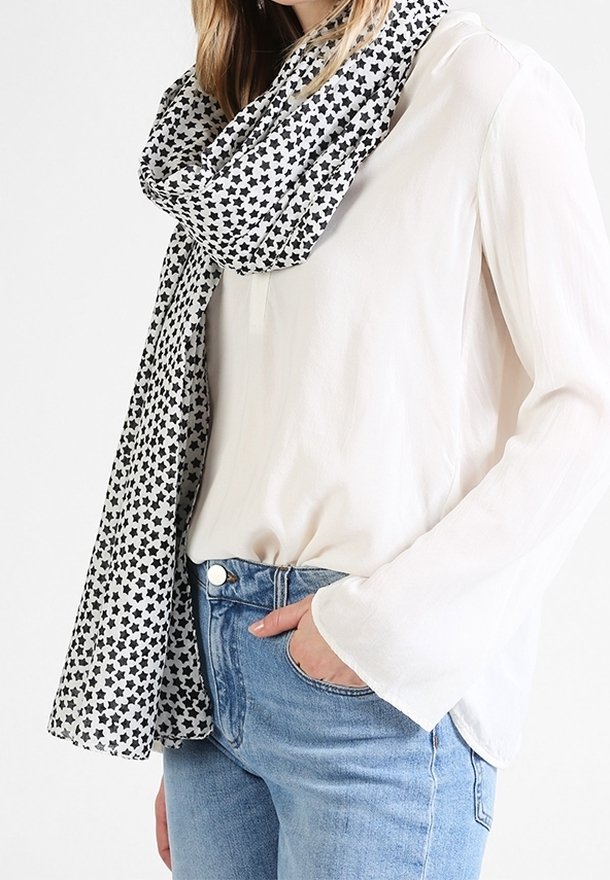 Femme portant un chemisier blanc à manches longues, un jean bleu et une écharpe blanche à motifs d'étoiles noires drapée autour du cou.