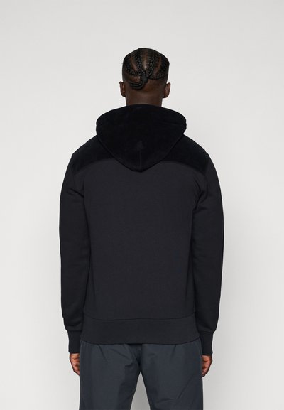 Un sweat à capuche noir avec une capuche noire texturée, présentant un tissu contrastant sur les épaules et un ourlet à côtes. Vue arrière affichée.