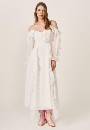 Mujer con un vestido largo blanco con hombros descubiertos, volantes y mangas transparentes, combinado con botas vaqueras metálicas rosas.