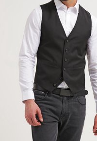 Schwarze maßgeschneiderte Weste mit sechs Knöpfen, ausgestattet mit zwei Fronttaschen, getragen über einem weißen Hemd. Kombiniert mit dunklen Jeans und einem schwarzen Gürtel.