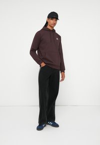 Jaunas vyras vilkintis tamsiai rudą Adidas džemperį su kapišonu, juodas džinsus, mėlynas Adidas sportines batus ir juodą kepurę, stovintis prieš baltą foną.