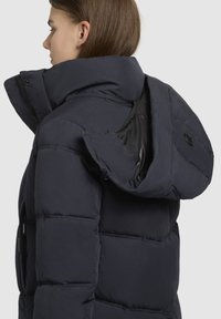 Marineblå pufferjakke med høj krave, lynlåslukning foran, stor hætte og udtalt quiltet tekstur. Har en skjult inderlomme.