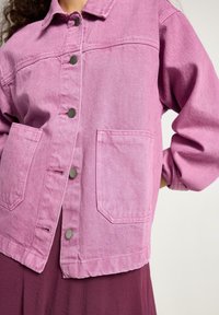 Růžová džínová bunda s límcem, zapínáním na knoflíky vpředu, dvěma náprsními kapsami a volnými rukávy. Texturovaná tkanina s jemným povrchem.