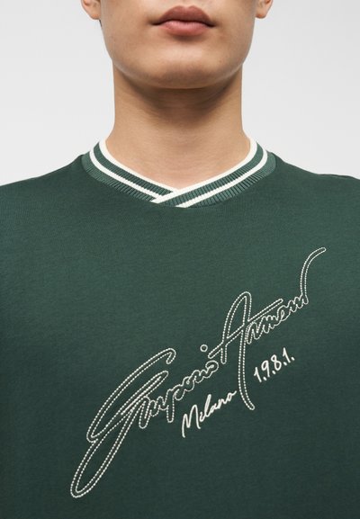 Πράσινη βαμβακερή μπλούζα με V-λαιμόκοψη, που διαθέτει αντίθετο λευκό ριμπ γιακά και διακοσμητικές λευκές ραφές με το όνομα της μάρκας και την ημερομηνία.