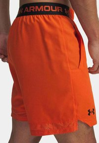 Pantaloni sportivi arancioni con una cintura elastica nera con la scritta "ARMOUR". Include tasche laterali e una rifinitura in rete all'orlo.