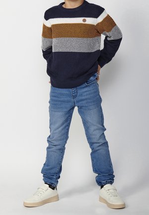 L'enfant se tient debout, les mains sur les hanches, portant un pull rayé bleu marine, blanc et marron, un jean bleu, des chaussettes noires et des baskets blanches.