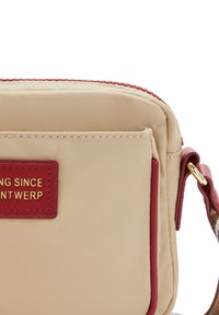Beige nylon crossover taske med røde detaljer, featuring et guldpræget mærke og en lynlåslukning. Glat tekstur, kompakt rektangulært design.