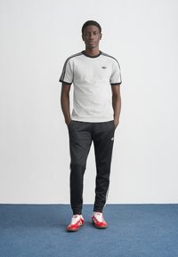 Siva majica s črnim robom in tremi črnimi črtami na rokavih, kombinirana s črnimi trenirkami s belimi stranskimi črtami. Rdeči športni copati.