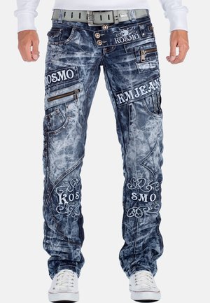 Mann trägt blaue Jeans im Acid-Wash-Stil mit mehreren aufgestickten Patches, Reißverschlüssen und einem grauen Gürtel, kombiniert mit weißen Turnschuhen und einem weißen Langarmshirt.