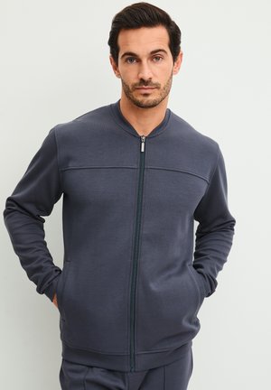 Homme aux cheveux courts et foncés portant une veste zippée bleu foncé, les mains dans les poches, devant un fond clair uni.