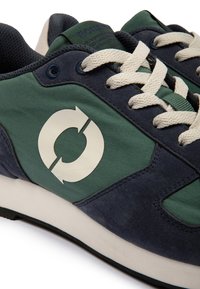 Baskets vertes et bleu marine avec lacets blancs et logo blanc circulaire sur le côté, montrant un gros plan des détails de la tige et de la semelle intermédiaire.